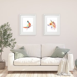 May include: Two framed watercolor art prints. One features a colorful bird perched on a branch, the other a fox with a unicorn horn. The prints are displayed above a white sofa with green pillows, creating a vibrant and artistic home decor scene.