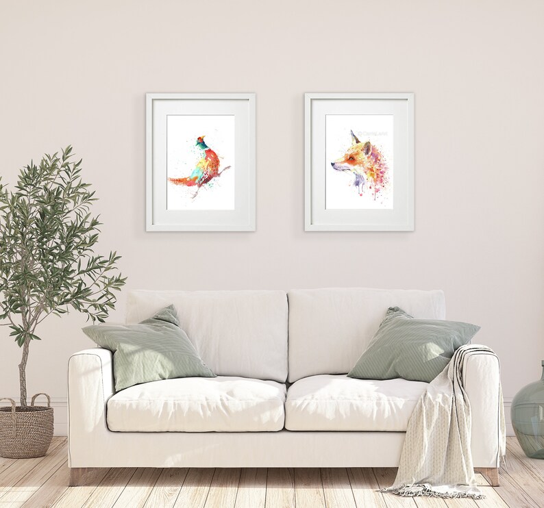 May include: Two framed watercolor art prints. One features a colorful bird perched on a branch, the other a fox with a unicorn horn. The prints are displayed above a white sofa with green pillows, creating a vibrant and artistic home decor scene.