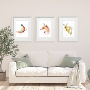 May include: Three framed watercolor animal prints. The prints feature a pheasant, a fox, and a rabbit, each rendered in vibrant splashes of color. The artwork is displayed above a white sofa with green pillows and a blanket, creating a cozy, artistic interior.
