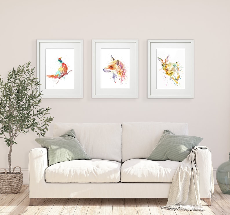 May include: Three framed watercolor animal prints. The prints feature a pheasant, a fox, and a rabbit, each rendered in vibrant splashes of color. The artwork is displayed above a white sofa with green pillows and a blanket, creating a cozy, artistic interior.