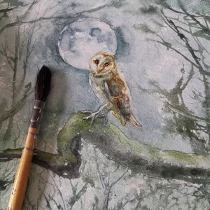 May include: Watercolor painting of a barn owl perched on a mossy branch, set against a backdrop of a full moon and silhouetted trees. The owl has brown and white feathers. A paintbrush is in the foreground.
