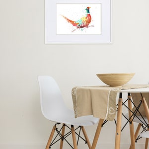 May include: A framed watercolor print of a pheasant in vibrant colors, including red, blue, and green. The artwork is displayed on a white wall above a table with a beige tablecloth and a wooden bowl.