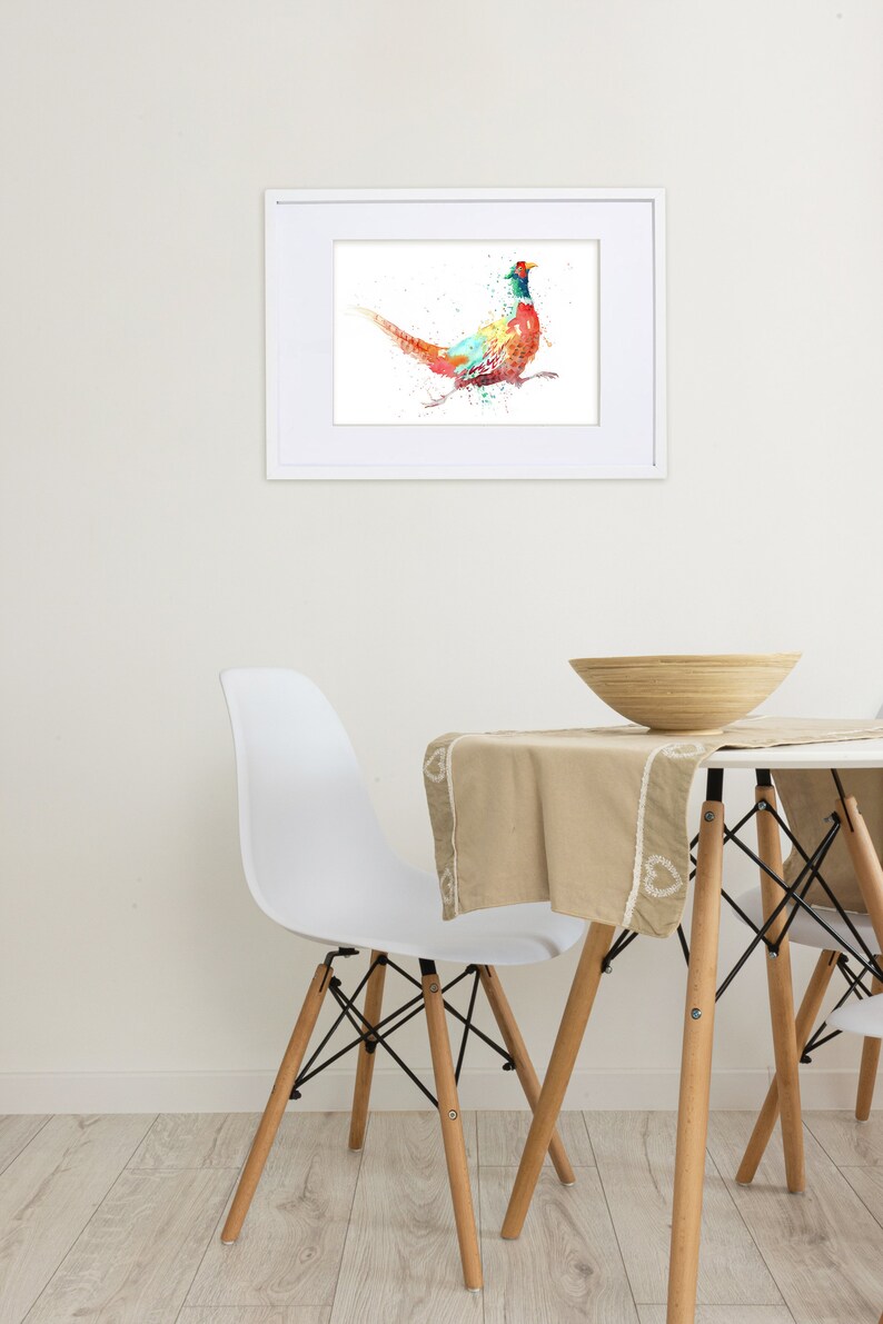 May include: A framed watercolor print of a pheasant in vibrant colors, including red, blue, and green. The artwork is displayed on a white wall above a table with a beige tablecloth and a wooden bowl.