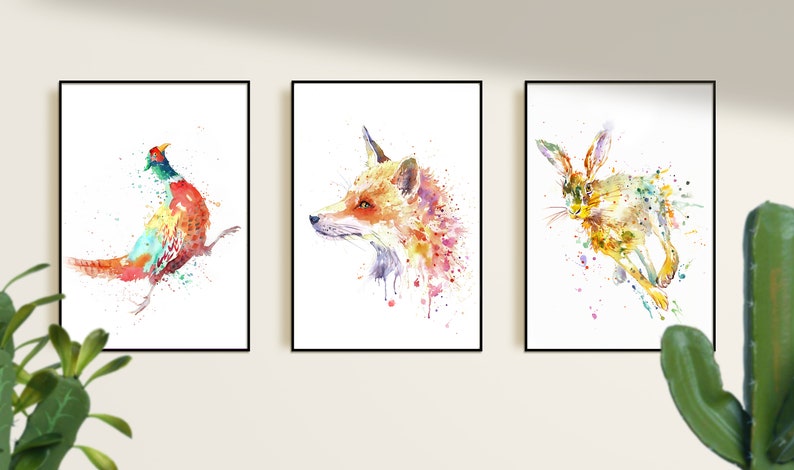 May include: Three framed watercolor prints featuring wildlife. The prints depict a pheasant, a fox, and a hare, each rendered in a vibrant, colorful, and abstract style. The artworks are framed in black and mounted on a white background.