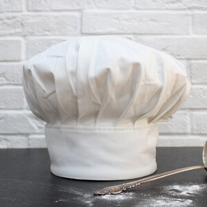 Puede incluir: Un gorro de chef blanco se encuentra sobre una superficie negra junto a un cuchar&oacute;n de plata. El gorro es alto y redondo con un dise&ntilde;o plisado. El cuchar&oacute;n tiene un mango largo y un taz&oacute;n redondo.