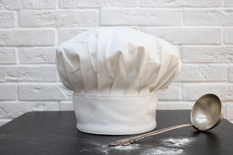 Puede incluir: Un gorro de chef blanco se encuentra sobre una superficie negra junto a un cuchar&oacute;n de plata. El gorro es alto y redondo con un dise&ntilde;o plisado. El cuchar&oacute;n tiene un mango largo y un taz&oacute;n redondo.