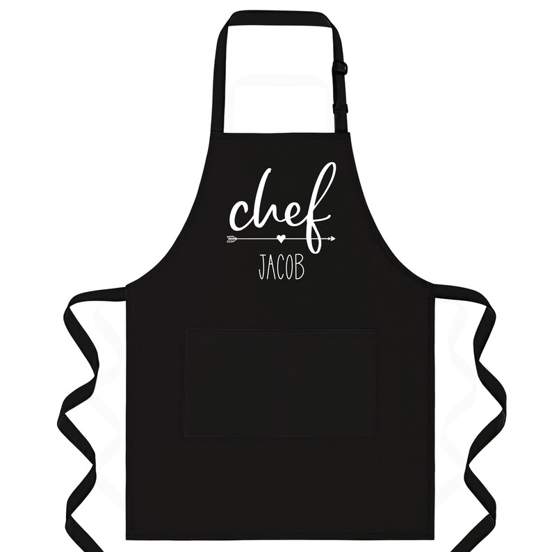 Puede incluir: Delantal de chef negro con un bolsillo blanco y el texto "chef" con una flecha y un coraz&oacute;n sobre el nombre "Jacob".