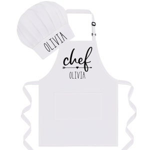 Puede incluir: Delantal de chef blanco con un bolsillo negro y una flecha negra con un coraz&oacute;n y el texto "chef Olivia". Se incluye un gorro de chef blanco con el texto "Olivia".