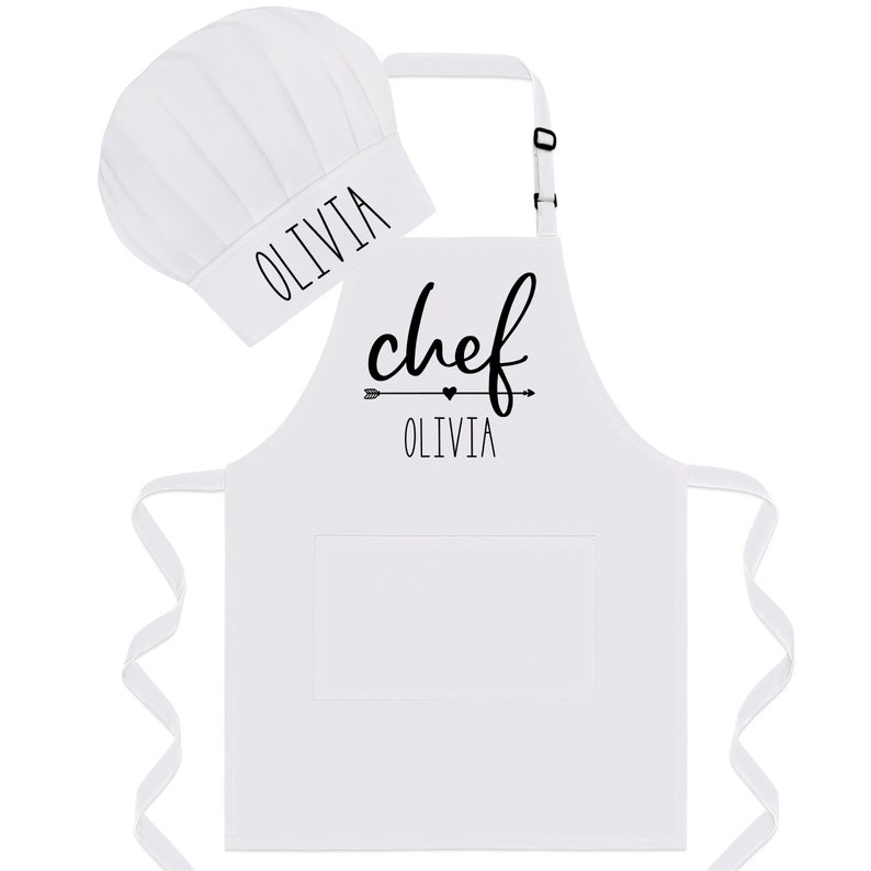 Puede incluir: Delantal de chef blanco con un bolsillo negro y una flecha negra con un coraz&oacute;n y el texto "chef Olivia". Se incluye un gorro de chef blanco con el texto "Olivia".
