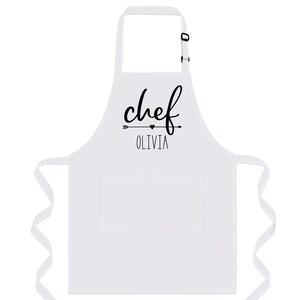 Puede incluir: Delantal de cocina blanco con una impresi&oacute;n negra que dice "chef Olivia" con una flecha y un coraz&oacute;n.