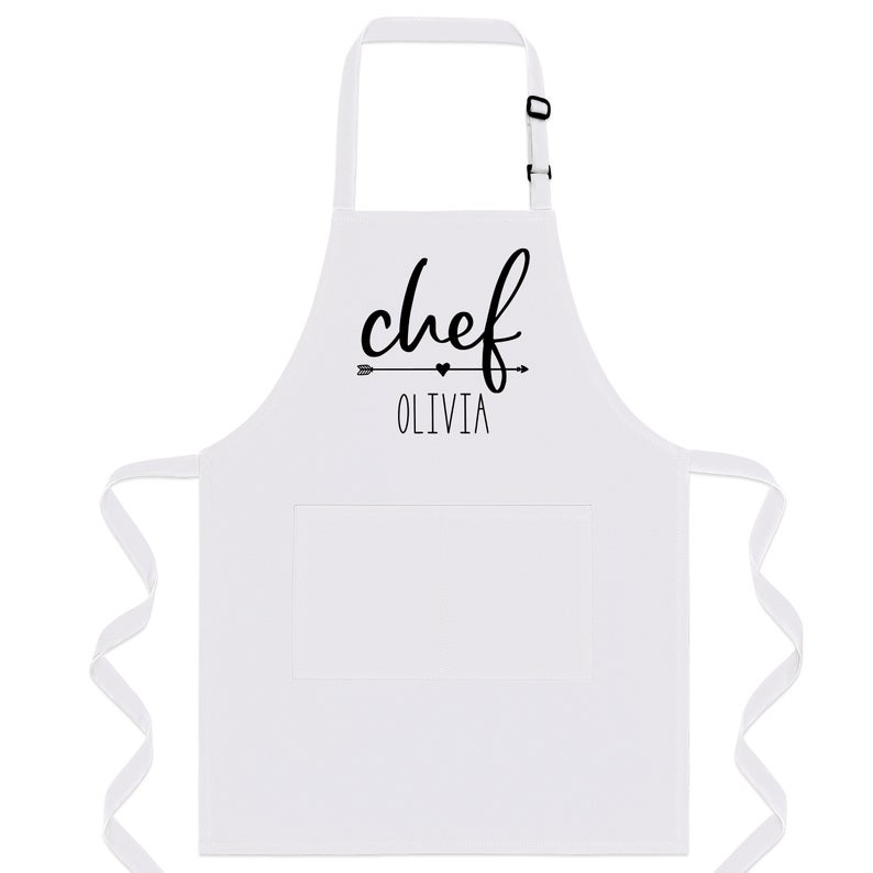 Puede incluir: Delantal de cocina blanco con una impresi&oacute;n negra que dice "chef Olivia" con una flecha y un coraz&oacute;n.