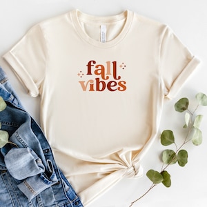 Peut inclure: Un t-shirt beige avec le texte "fall vibes" en lettres brunes, orange et roses. Le t-shirt est plié et posé sur une surface blanche avec une veste en jean et une brindille de verdure.