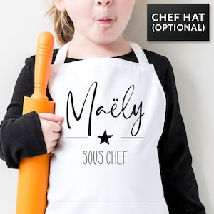 May include: White apron with black text that says "Maëly Sous Chef" with a black star in between the words. A child is wearing the apron and holding a rolling pin.