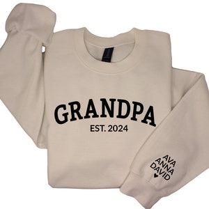 Peut inclure: Un sweat-shirt crème avec le texte "GRANDPA EST. 2024" imprimé sur le devant. La manche gauche porte les noms "AVA ANNA DAVID" imprimés avec un symbole de cœur.
