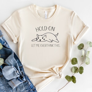 May include: A cream-coloured t-shirt featuring the text "HOLD ON LET ME OVERTHINK THIS..." and a black cat illustration. The shirt is folded at the bottom and is paired with a denim jacket and a sprig of greenery.