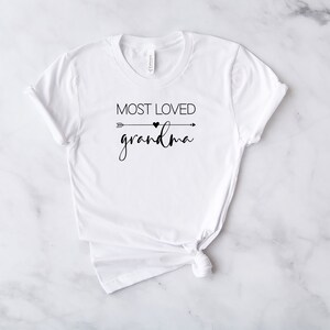 May include: White t-shirt with black text that reads "Most Loved Grandma" with a small heart and arrow graphic.