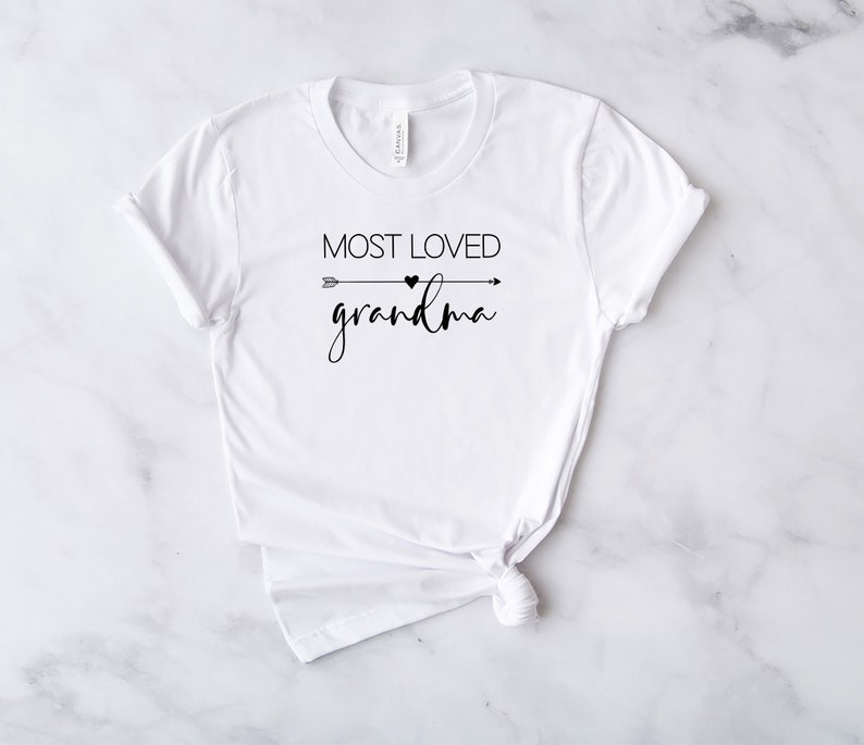 May include: White t-shirt with black text that reads "Most Loved Grandma" with a small heart and arrow graphic.