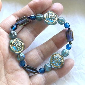 May include: A beaded bracelet featuring a mix of blue and teal beads. The bracelet includes faceted, round, and rectangular beads, with three decorative flower-shaped beads. The bracelet is held in a hand.