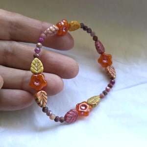 May include: A beaded bracelet featuring orange flower and leaf-shaped beads interspersed with small, round beads in shades of pink, brown, and gold. The bracelet is held in a hand, showcasing its autumn-inspired color palette and design.