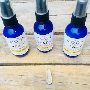Puede incluir: Tres botellas de spray de vidrio azul con tapas negras y etiquetas blancas. Las etiquetas dicen "The Moon & The Spade" y tienen texto diferente debajo. Las botellas están sobre una superficie de madera con un cristal de cuarzo transparente en el centro.
