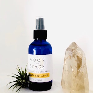 May include: A blue glass bottle with a black spray top and a white label that reads "The Moon & The Spade Energetic Boundaries Aura Protection 2 fl oz / 60 ml". The bottle is sitting on a white surface next to a clear quartz crystal.