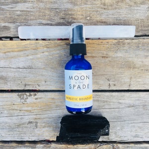 May include: A blue glass bottle with a black spray top and white label. The label reads "The Moon & The Spade Energetic Boundaries 2 fl oz / 60 ml". The bottle is sitting on a wooden surface with a white and black crystal next to it.