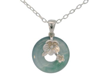 Puede incluir: Collar de plata con un colgante de jade verde pálido de forma circular, con un diseño floral. El colgante tiene un agujero central y está adornado con dos flores de plata. La cadena también es de plata.