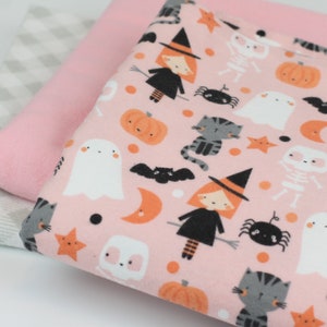 May include: Three fabric pieces: a pink solid, a gray and white checkered, and a pink Halloween-themed print. The print features witches, ghosts, pumpkins, cats, bats, and stars on a pink background. The fabrics are stacked.