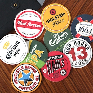 Könnte beinhalten: Eine Sammlung runder, bunter Bieruntersetzer mit verschiedenen Markennamen und Logos. Die Untersetzer haben Designs in Grün, Rot, Gelb, Weiß und Schwarz. Einige der Marken sind Corona, Red Arrow, Holsten, Camden und Newcastle Brown Ale.