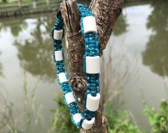 May include: A blue and gold braided collar with white rectangular beads. The collar is displayed on a tree branch against a blurred background of water and greenery. The collar is likely for a pet.