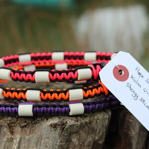 May include: Three colorful dog collars with white beads. The collars are pink, orange, and purple. The collars are on a wooden surface with a white tag that says "Hope 36cm Rose/noir Kalaa 41cm violet/noir Shenzy 38cm orange/noir 100% fait avec  love"