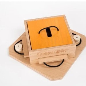 May include: A wooden dog puzzle with a light brown base and a square box with an orange top. The box has a black handle and a hole. The words "Intelligent Dogs" are printed on the side. Two white cylinders are attached to the base.
