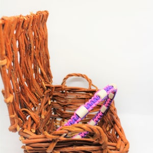 May include: A woven wicker basket with a purple and pink braided bracelet with white beads inside.