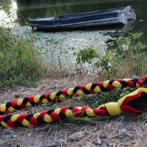 Può includere: Un giocattolo per cani intrecciato con tessuto nero, giallo e rosso. Il giocattolo è sdraiato a terra vicino a uno specchio d'acqua.