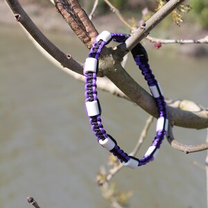 May include: A purple and black paracord bracelet with white beads. The bracelet is hanging from a tree branch.