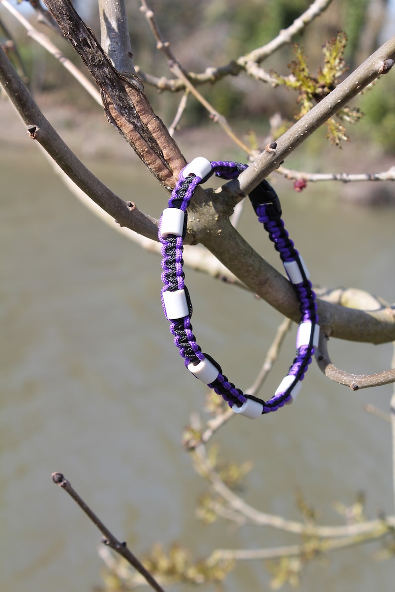 May include: A purple and black paracord bracelet with white beads. The bracelet is hanging from a tree branch.