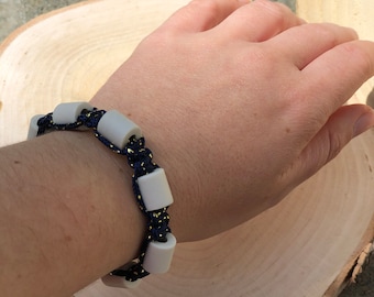 May include: A handmade bracelet featuring white cylindrical beads strung on a navy blue cord with gold accents. The bracelet is displayed on a person's wrist, showcasing its design and craftsmanship. The jewelry is suitable for everyday wear.