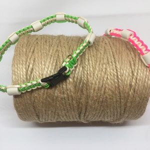 May include: Two colorful paracord collars with white beads and black buckles. The collars are green and pink and are wrapped around a roll of twine.