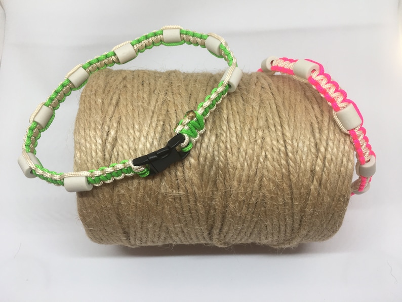May include: Two colorful paracord collars with white beads and black buckles. The collars are green and pink and are wrapped around a roll of twine.