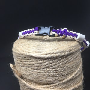 May include: A purple and white braided bracelet with a black plastic clasp. The bracelet is on a roll of twine.