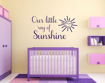 Peut inclure: Décoration de chambre d'enfant en violet et blanc avec un lit bébé et un autocollant mural qui dit "Notre petit rayon de soleil" avec un graphique de soleil souriant.