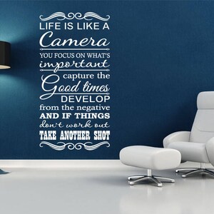 Op de afbeelding: Witte vinyl wanddecoratie met een citaat over het leven dat als een camera is. De quote luidt: "Het leven is als een camera. Je focust op wat belangrijk is, je legt de goede tijden vast, je ontwikkelt vanuit het negatief, en als dingen niet lukken, maak je gewoon een nieuwe foto."