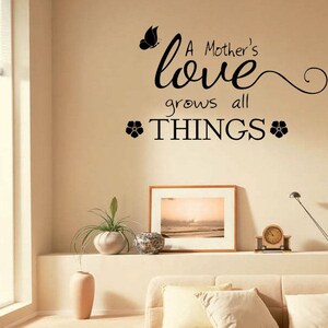 Op de afbeelding: Een zwart-witte muursticker met de tekst "A Mother's love grows all THINGS" met een vlinder en twee bloemmotieven.