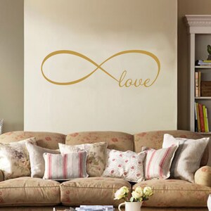 Könnte beinhalten: Ein goldenes Unendlichkeitszeichen mit dem Wort "love" darin. Das Symbol befindet sich an einer beige Wand über einem braunen Sofa mit floralen Kissen.