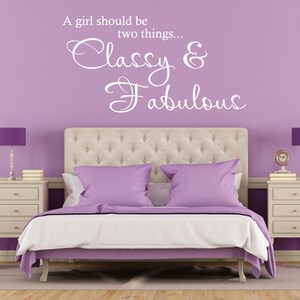 Op de afbeelding: Een paarse slaapkamer met een wit gestoffeerd hoofdeinde en een wit citaat op de muur dat luidt: "Een meisje moet twee dingen zijn... Classy & Fabulous".