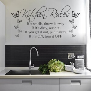 Puede incluir: Una cocina blanca con un salpicadero de azulejos negros y un fregadero de acero inoxidable. La pared tiene una calcomanía de vinilo negro con el texto "Kitchen Rules" y una lista de reglas. El texto está rodeado de siluetas de mariposas.