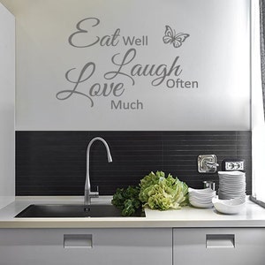 May include: A kitchen with a grey tiled backsplash and a white worktop. The wall has a grey vinyl decal with the text "Eat Well Love Much Laugh Often" and a butterfly graphic.