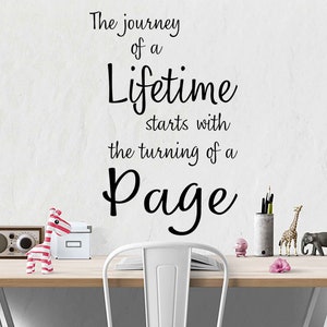 Könnte beinhalten: Ein weißer Schreibtisch mit einer Holzplatte und einem weißen Stuhl. Die Wand hinter dem Schreibtisch hat einen schwarzen Vinyl-Aufkleber mit dem Zitat "The journey of a lifetime starts with the turning of a page".