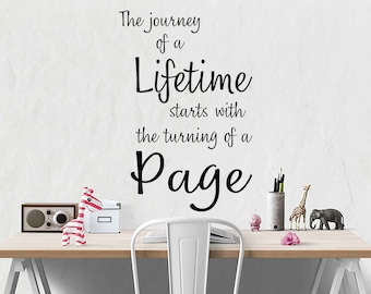 May include: A white desk with a wooden top and a white chair. The wall behind the desk has a black vinyl decal with the quote "The journey of a lifetime starts with the turning of a page".