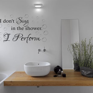 Puede incluir: Un baño con un lavabo blanco, una encimera de madera y un espejo. La pared tiene una calcomanía de vinilo negro que dice "No canto en la ducha. Actúo."